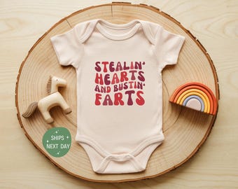 May include: A light pink baby onesie with the text "Stealin' Hearts and Bustin' Farts" in red and maroon lettering. The onesie is on a wooden slice with a wooden horse and a rainbow.