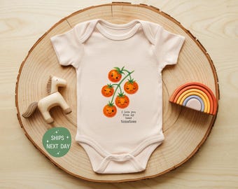 May include: A light pink baby onesie with a graphic of smiling orange tomatoes on a green vine. The text "I love you from my head tomatoes" is below the tomatoes. The onesie is on a wooden slice with a wooden horse and rainbow.