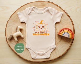 May include: A light beige baby onesie with a pumpkin graphic and the text "MY FIRST HALLOWEEN". The design includes bats and stars. A wooden horse and a rainbow are next to the onesie.