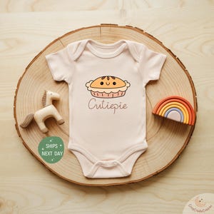 May include: A light pink baby onesie with a cartoon pie graphic and the word "Cutiepie" in brown script. The onesie is displayed on a wooden slice with a wooden horse and a rainbow-shaped toy.