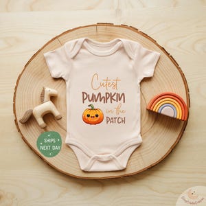 May include: A light beige baby onesie with the text "Cutest PUMPKIN in the PATCH" and a cartoon pumpkin graphic. The onesie is on a wooden slice with a wooden horse and a small rainbow. A fall-themed baby outfit.