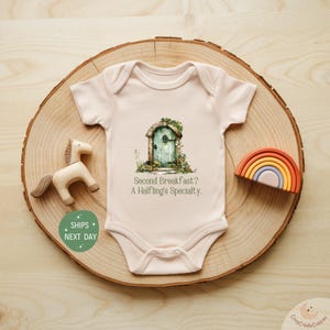 May include: A light pink baby onesie with a whimsical illustration of a green door with the text "Second Breakfast? A Halfling's Specialty." A wooden horse and a rainbow are nearby.