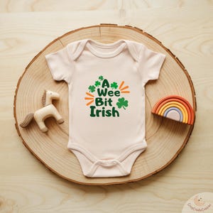 May include: A light pink baby onesie with the text "A Wee Bit Irish" in green and orange. The onesie is displayed on a wooden slice with a wooden horse and a rainbow toy.