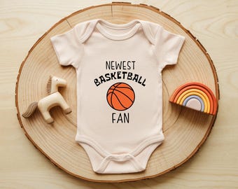 May include: A light pink baby onesie with the text "Newest Basketball Fan" and a basketball graphic. The onesie is displayed on a wooden slice with a wooden horse and a rainbow-shaped toy.