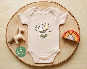 May include: A light pink baby onesie with a panda bear graphic and the text "Mommy's Little Panda." The onesie is displayed on a wooden slice with a wooden horse and a rainbow-shaped toy.