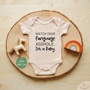 May include: A light pink baby bodysuit with the text "WATCH YOUR language ASSHOLE. I'm a Baby." is displayed on a wooden slice. A wooden horse and a rainbow-shaped toy are next to the bodysuit.