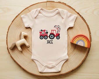 May include: A light pink baby onesie with a red tractor design pulling a wagon filled with red and black hearts. The name "JACE" is printed below the tractor. A wooden horse and rainbow are next to the onesie.