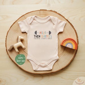 May include: A light pink baby onesie with the text "MILK THEN BURPEES TRAINING STARTS EARLY" in a playful font. The onesie is displayed on a wooden slice with a wooden horse and a rainbow-shaped toy.