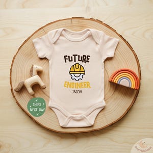 May include: A light beige baby onesie with the text "FUTURE ENGINEER" and a yellow hard hat graphic. The name "JASON" is printed below. A wooden horse and rainbow are on a wooden slice.