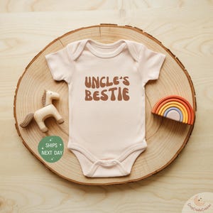 May include: A light pink baby onesie with the text "Uncle's Bestie" in brown letters with a retro font style. The onesie is on a wooden surface with a wooden toy horse and a rainbow-colored wooden toy.