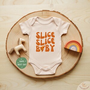 May include: A light pink baby onesie with the words "SLICE SLICE BABY" in orange lettering and a pizza slice graphic. The onesie is displayed on a wooden slice with a wooden horse and rainbow.