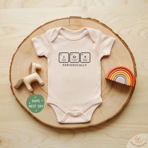 May include: A light pink baby onesie with the word "PERIODICALLY" printed below three boxes with chemical element symbols. A wooden horse and a rainbow-shaped toy are next to the onesie.
