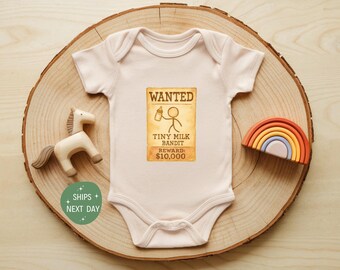 May include: A light pink baby onesie with a "Wanted" poster graphic that reads "Tiny Milk Bandit" and "Reward: $10,000". The onesie is on a wooden slice with a wooden horse and a rainbow.