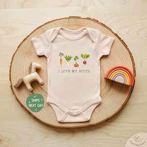 May include: A light pink baby onesie with a watercolor illustration of various root vegetables, including carrots, radishes, and potatoes. The text "I LOVE MY ROOTS" is printed below the vegetables. The onesie is displayed on a wooden slice with a toy horse and rainbow.