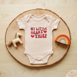 May include: A light pink baby onesie with the text "MY LITTLE HEART THIEF" in shades of red. A wooden toy horse and a rainbow-shaped toy are next to the onesie on a wooden slice.