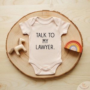 May include: A light pink baby onesie with the text "TALK TO MY LAWYER" in black. The onesie is on a wooden slice with a wooden horse and a rainbow-shaped toy.