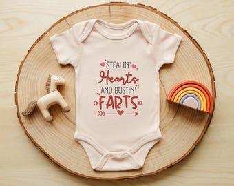 May include: A light pink baby onesie with the text "Stealin' Hearts and bustin' Farts" in red. The onesie is on a wooden slice with a wooden horse and a small rainbow. A cute and funny baby outfit.