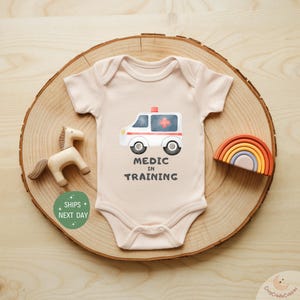 May include: A light pink baby onesie with a watercolor ambulance graphic and the text "MEDIC IN TRAINING". The onesie is displayed on a wooden slice with a wooden horse and a rainbow toy.