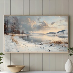 Könnte beinhalten: Ein gerahmtes Landschaftsgemälde zeigt eine verschneite Seeuferlandschaft mit kahlen Bäumen und einem bewölkten Himmel. Das Kunstwerk wird über einem weißen Schrank mit einer Holzschale und dekorativen Vasen ausgestellt.