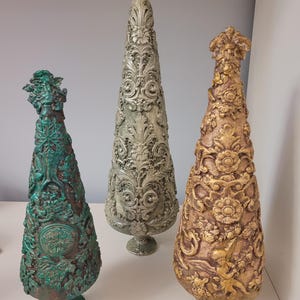 May include: Three decorative, cone-shaped ornaments in green, silver, and gold. Each has an embossed design and sits on a pedestal base. The tallest has a bow at the top. These are likely for home decor, with detailed, ornate patterns.