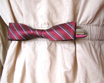 May include: A red and blue striped bow tie with a silver metal clasp is attached to a beige fabric garment.