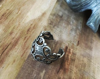 May include: An antique silver-colored adjustable ring with an openwork design. The ring features floral and geometric patterns, including a central starburst design. The ring is displayed on a wooden surface.