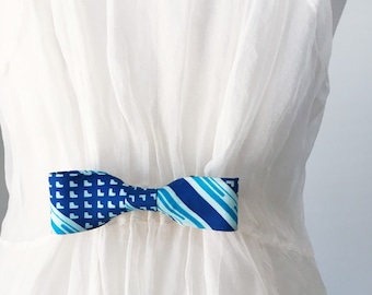 May include: A blue bow tie with a white dress. The bow tie features two distinct patterns: one side with a repeating square design and the other with blue and light blue stripes. The bow tie is a stylish accessory.