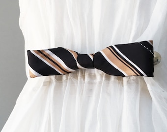 May include: A black, tan, and white striped bow tie, knotted in the center, is attached to a white garment. The bow tie is the focal point, with its diagonal stripes creating a striking contrast against the white fabric. The bow tie is approximately 5 inches wide.