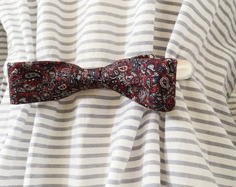 May include: A red and blue paisley bow tie with a white clip on a striped background.