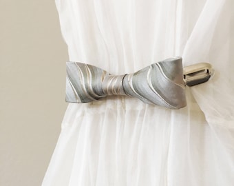 May include: A green and gold patterned bow tie curtain tieback with a silver metal clip.