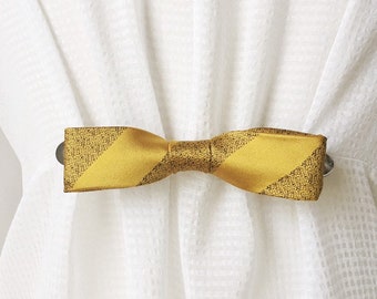 May include: A gold and black striped bow tie curtain tieback. The tieback is made of fabric and has a metal clip.