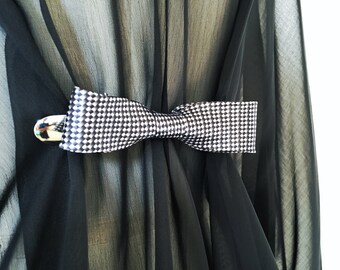 May include: A black and white patterned bow tie clip, made of fabric, with a silver metal clasp. The clip is attached to a black sheer fabric.