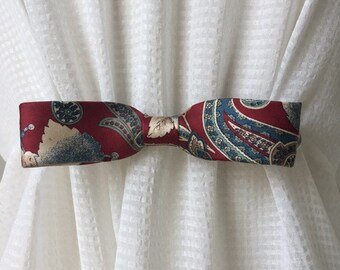 May include: A red, blue, and cream patterned fabric bow tie curtain tieback. The tieback is tied in a bow shape.