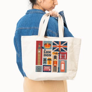 May include: Cream-colored tote bag featuring a colorful London-themed design. The design includes iconic landmarks like Big Ben, a red telephone booth, and the Union Jack flag. The bag is held by a person wearing a denim jacket.