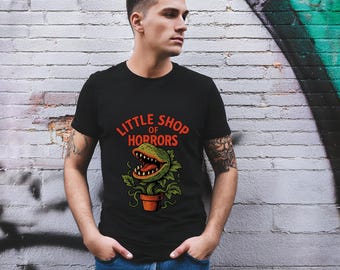 Puede incluir: Camiseta negra con un gráfico de una planta carnívora en una maceta y el texto "Little Shop of Horrors" en naranja. La planta tiene una gran boca abierta con dientes y una lengua roja. La camiseta es de manga corta.