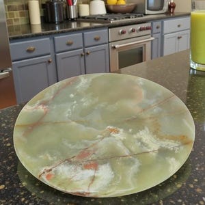 May include: A round, light green onyx serving plate with reddish-brown veining. The plate has a polished surface and is set on a dark countertop. The plate is approximately 12 inches in diameter. Kitchenware.