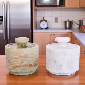 May include: Two stone jars with lids sit on a wooden surface. One jar is green and brown, the other is white. The jars have a cylindrical shape with a rounded base and a knob on the lid. The background shows a kitchen setting.