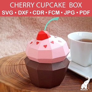 May include: A papercraft cherry cupcake box with a red cherry on top and heart accents. The box is pink and brown, with the text "CHERRY CUPCAKE BOX" at the top. A cup of tea sits next to the box.