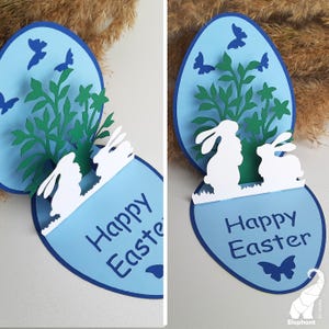May include: A blue and white pop-up card with a 3D Easter scene. The card features two white bunnies, green foliage, and blue butterflies. The card reads "Happy Easter".
