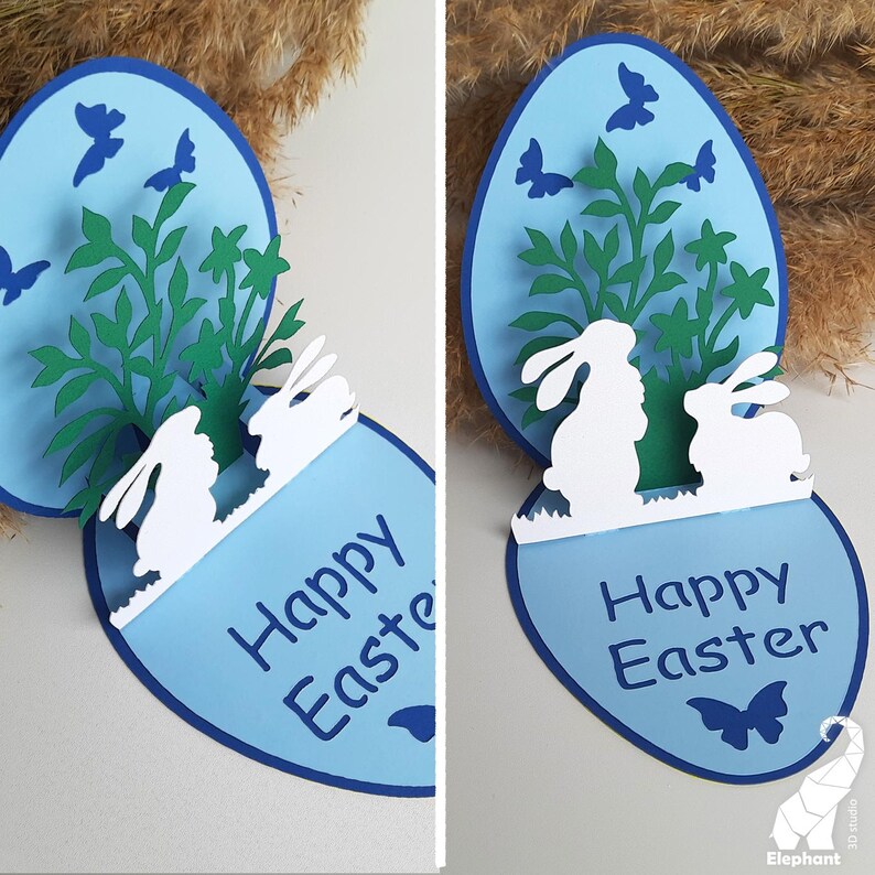 May include: A blue and white pop-up card with a 3D Easter scene. The card features two white bunnies, green foliage, and blue butterflies. The card reads "Happy Easter".