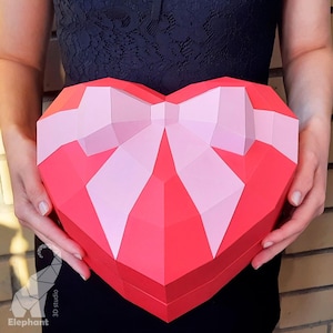 May include: A red heart-shaped box with a pink bow. The box is made of paper and has a 3D design.
