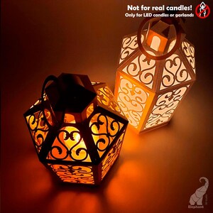May include: Two decorative paper lanterns with intricate cut-out designs. The lanterns are hexagonal and have a warm, inviting glow from the LED lights inside. The text "Not for real candles! Only for LED candles or garlands" is visible.
