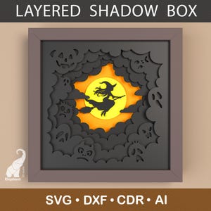 May include: A layered shadow box with a Halloween theme. The artwork features a witch on a broomstick silhouetted against a yellow sun, surrounded by dark gray clouds, ghosts, and jack-o'-lanterns. The frame is a muted gray color. Text at the top reads "LAYERED SHADOW BOX".