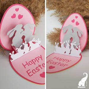 May include: A pink and white pop-up card with a 3D design of bunnies inside an egg. The card reads "Happy Easter".