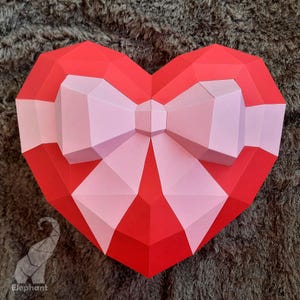 May include: A red and pink paper heart with a large bow in the center. The heart is made of folded paper and has a three-dimensional effect.