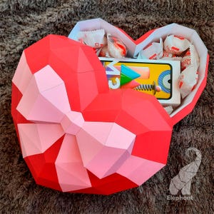 May include: A red and pink paper heart-shaped box with a bow. The box is open and filled with candy and a small electronic device.
