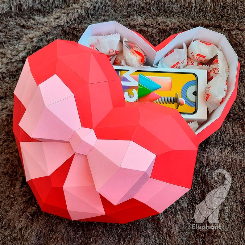 May include: A red and pink paper heart-shaped box with a bow. The box is open and filled with candy and a small electronic device.