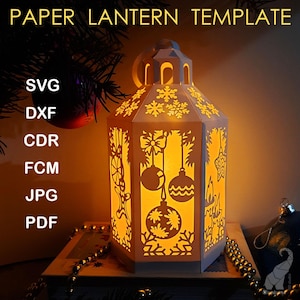 Op de afbeelding: Een verlichte papieren lantaarn sjabloon met een feestelijk ontwerp. De witte lantaarn heeft uitgesneden patronen van sneeuwvlokken, kerstballen en strikken. De tekst "PAPER LANTERN TEMPLATE" staat bovenaan. Bestandsformaten zijn SVG, DXF, CDR, FCM, JPG en PDF.