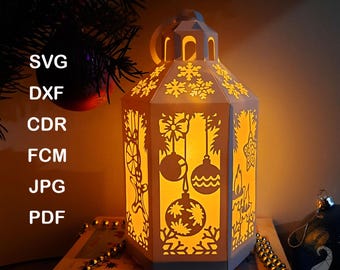 May include: A lit paper lantern template with a festive design. The lantern is white with cut-out patterns of snowflakes, ornaments, and bows. The text "PAPER LANTERN TEMPLATE" is at the top. File formats include SVG, DXF, CDR, FCM, JPG, and PDF.