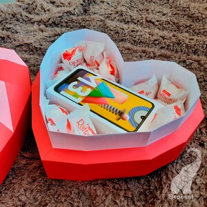 May include: A red heart-shaped box with a white interior, filled with small white chocolate candies and a yellow and white game card. The box is made of paper and has a folded design.